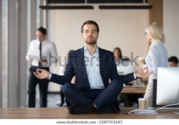 Mindful calm businessman in suit meditating at\
office sitting in lotus position on work desk, successful ceo\
executive doing yoga exercise at workplace, peace of mind, no\
stress free relief\
concept