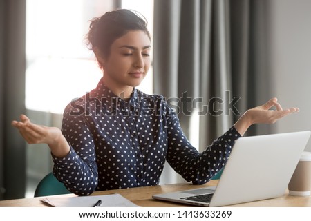 Mindful calm indian young business woman meditate at work desk with eyes closed, healthy hindu girl take break relax in office doing yoga at workplace feel balance no stress and peace of mind concept
