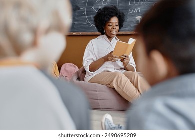 Mindful female teacher of Black ethnicity in bean bag chair against blackboard reading aloud gripping story to kids sitting around on classroom floor, copy space - Powered by Shutterstock