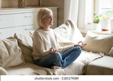 Mindful Healthy Mature Woman Practicing Meditation At Home, Calm Senior Middle Aged Lady Sitting On Couch In Lotus Pose Doing Yoga For Mental Balance Breathing Air Relaxing On Stress Free Weekend