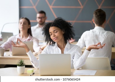 Mindful millennial african woman meditating at workplace with colleagues, calm black employee or student doing yoga exercises feeling zen enjoying no stress free relief, corporate meditation concept