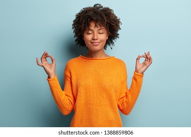 Mindful peaceful Afro American woman meditates indoor, keeps hands in mudra gesture, has eyes closed, tries to relax after long hours of working, holds fingers in yoga sign, isolated on blue wall