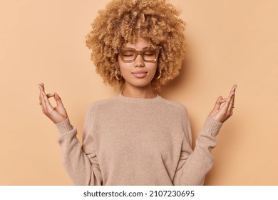 Mindful People. Calm Young Woman Breathes Peaceful Meditates With Closed Eyes Practices Yoga Stands Relaxed Wears Jumper And Spectacles Isolated Over Beige Background. Zen And Patience Concept