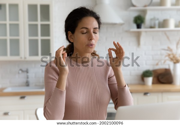 Mindful young woman breathing out with closed\
eyes, calming down in stressful situation, working on computer in\
modern kitchen. Millennial hispanic lady managing stress, practice\
yoga at home office.