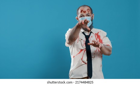 Mindless office zombie with covid protection mask and bloody wounds standing on blue background. Creepy scary brain-eating monster with protective facemask and deep scars growling at camera.