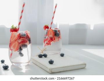 Mineral Infused Water In A Glass With Strawberry And Blueberry And Paper Straws On The White Wood Board With White Background And Copy Space