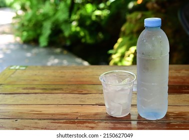 Mineral Water Is Packed In Clear Plastic Bottle, The Blue Cap Has Condensation Due To The Cold. Plastic Glasses Filled With Ice Were Placed Side By Side On Wooden Table With Blurred Natural Backgroud.