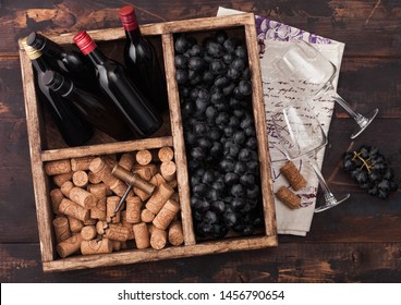 Mini bottles of red wine and empty glasses with dark grapes with corks and corkscrew inside vintage wooden box on dark wooden background with linen towel.