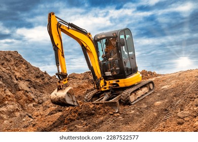 Mini excavator at the construction site on the edge of a pit against a cloudy blue sky. Compact construction equipment for earthworks. An indispensable assistant for earthworks