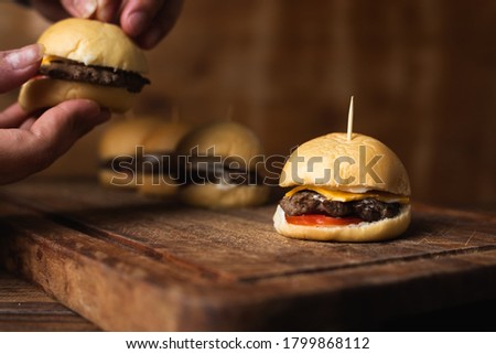 Mini homemade burger with cheddar cheese, tomato and mayonnaise to eat them as snacks. Hands grabbing a burger in the background. Horizontal image with space for text.