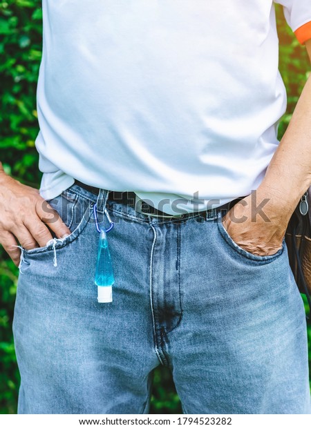 Mini portable alcohol gel bottle to kill Corona\
Virus(Covid-19) hanging on belt loop of man\' s jeans with plants\
background. New normal lifestyle. Health care concept. Selective\
focus on alcohol gel