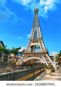 A mini replica of the Eiffel Tower located in a tropical outdoor park with a clear blue sky background. The structure is surrounded by decorative fences, lush green trees, and colorful architecture, 
