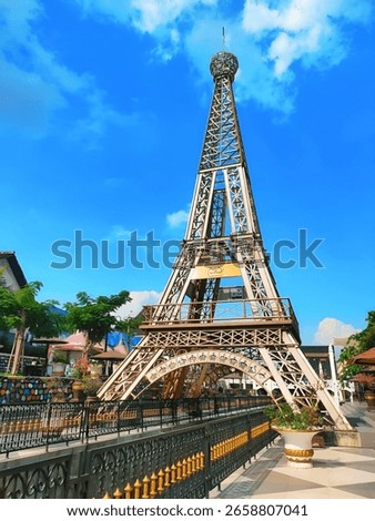 A mini replica of the Eiffel Tower located in a tropical outdoor park with a clear blue sky background. The structure is surrounded by decorative fences, lush green trees, and colorful architecture, 