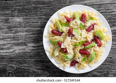 Mini Salami Bowtie Pasta Warm Salad With Avocado Slices And Basil Leaves Sprinkled With Grated Parmesan Cheese On White Plate On Dark Old Wooden Background, View From Above