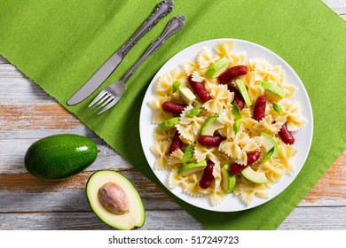 Mini Salami Bowtie Pasta Warm Salad With Avocado Slices And Basil Leaves On White Plate On Table Mat With Fork And Knife, Half Of Ripe Avocado On Old Wooden Background, View From Above