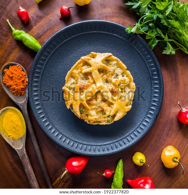 Mini tasty\
chicken pie on black plate with peppers, wooden spoon parsley,\
saffron and spicy paprika on wooden background. Top view of chicken\
pie with spices and\
vegetables.
