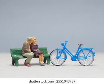 Mini toy of action figure at outdoor with blurred background. Toy photography concept design. Minifigure of old couple character sitting on the bench with blue bicycle. Romantic scene at garden. - Powered by Shutterstock