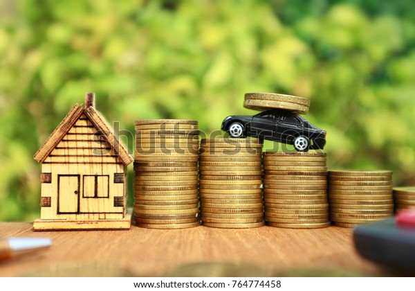 Miniature
black car carry Dollar Singapore coins and drives on rolls ladder
of gold money lead to wooden house,  calculator and pen on wood
table in blurred natural tree bright
light