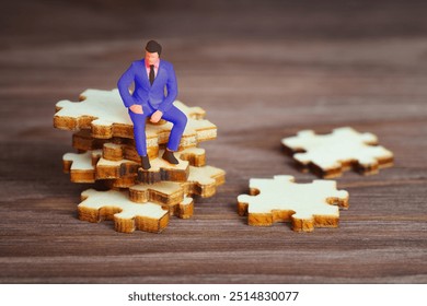 Miniature businessman figurine perched on stacked puzzle pieces on wooden surface, symbolizing problem-solving and decision-making. - Powered by Shutterstock