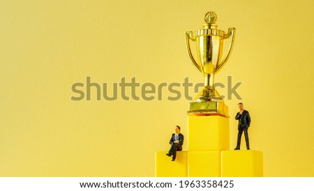 Miniature of businessman stands on podium ladder with golden trophy on yellow background