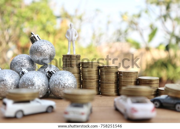 Miniature businessman stands on top of rolls\
ladder step of gold money to pole of Christmas glitter silver balls\
decorations and cars carry coins on wood table in blur natural tree\
bright sunlight
