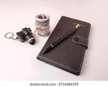 Miniature camera keychain, a roll of US currency tied with string, and a black leather notebook with a pen resting on it.