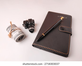 Miniature camera keychain, a roll of US currency tied with string, and a black leather notebook with a pen resting on it.
