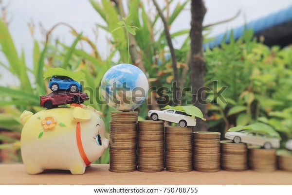 Miniature cars on piggy bank and vehicles carry\
green leaves to world globe on rolls ladder of gold coin money on\
wood table in blur natural\
tree