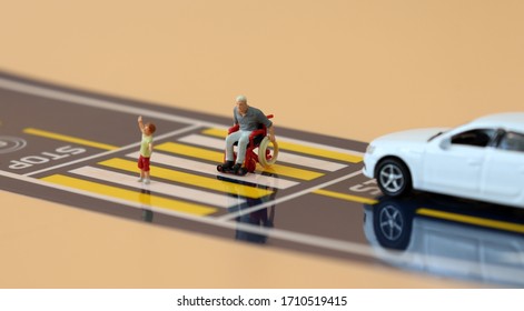 A Miniature Child With A Hand Raised And A Miniature Man  In A Wheelchair Crossing A Crosswalk.