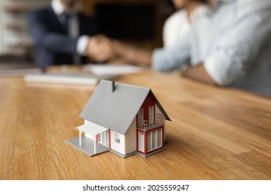 Miniature Of Cottage House On Table Close Up View, Happy African Family, Agency Or Bank Clients Handshake Real Estate Agent After Contract Signing, On Blurred Background. Tenancy And Mortgage Concept