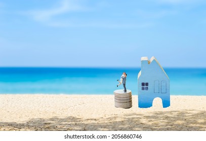 Miniature couple on stack of coin with blue wooden hosue model on the beach with space on blue sea background, summer house, investment and real estate business concept