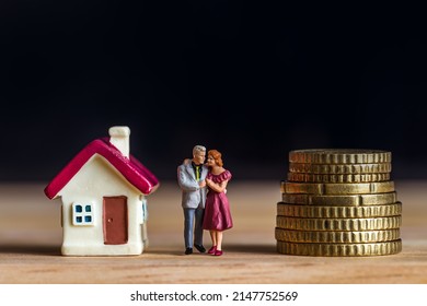 Miniature Couple With Tiny Home  And Stack Coins Over Blurred Dark Background.