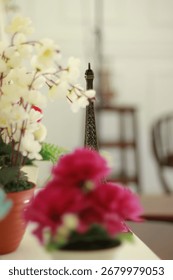 A miniature Eiffel Tower sits between two potted artificial flowers, one white and one pink, on a table indoors.