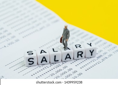 Miniature Figure Working Businessman, Office Guy Walking On Cube Block Building Word Salary On Bank Deposit Account Book On Yellow Background, Salary Man, Office Wage Or Employee Bank Deposit Concept.