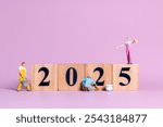 Miniature figures in overalls paint and construct the numbers "2025" on wooden blocks against a pastel pink background.