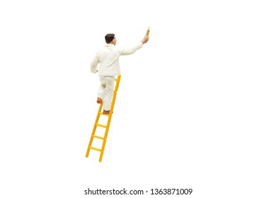 Miniature Figurine Character As Painter Standing On Wooden Ladder And Painting Wall With Paint Tools Isolated On White Background.