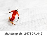 A miniature figurine of a skier dressed in vibrant winter gear. The skier is captured in motion, skiing across a white snowy landscape