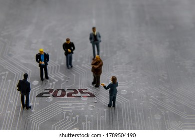 Miniature figurines posed as business people in a meeting over abstract futuristic circuit board with text year 2025