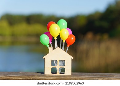 Miniature Flat House Shape With A Bunch Of Small Colorful Balloons Against A Beautiful Lake Landscape Background. Creative Moving In Party Concept.