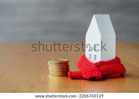 Miniature house in a red scarf with coins on wooden table. The concept of passive house heating. Thermal insulation of a building or dwelling. Energy crisis.