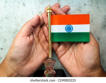 Miniature Indian National Flag On A Metal Stand With National Emblem 'Ashok Stambh' In A Hand On White Marble Background To Show Respect, Love, Care And Protection Concept For The Country.