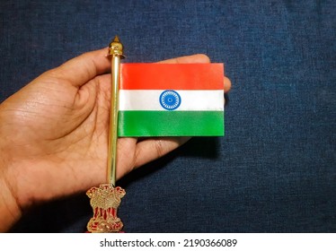 Miniature Indian National Flag On A Metal Stand With National Emblem 'Ashok Stambh' In A Hand On Blue Canvas Background To Show Respect, Love, Care And Protection Concept For The Country.