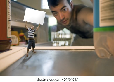 A Miniature Man Holding Up A Blank Sign Inside The Refrigerator.  Possible Text On The Sign Could Be WHATS FOR DINNER Or WE NEED FOOD.  Use Your Imagination!  Shallow Depth Of Field.