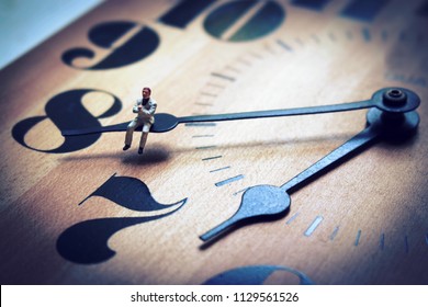 Miniature Man Sitting On The Hands Of A Clock. Concept Of Waiting Or Running Out Of Time. Work Or Business Deadline Is Ticking Away. Employee Or Boss Struggles With Time Management. 