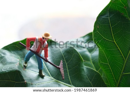 Miniature model, a farmer guy wearing a hat, blue pants, orange shirt, green boots, holding a rake. He is working on a green fiddle fig leaf