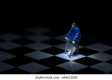 Miniature People: Badly Injured Small Figure Businessman Walking On Crutches With Chess Board And Black Background. Health And Insurance Concept.