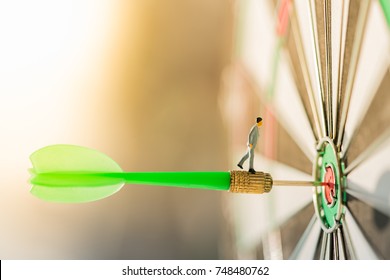 Miniature People: Small Business Man On Red Dart Arrow Hitting In The Target Center Of Dartboard With Modern City And Sunset Background. Target Business, Achieve And Victory Concept .