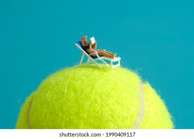 Miniature People Toy Figure Photography. Girl Relaxing Seat At Beach Chair While Reading Book Above Tennis Ball. Image Photo