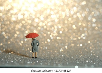 Miniature People Toy Figure Photography. A Lady Wearing Raincoat And Umbrella, Standing Enjoying Golden Yellow Bokeh Light Background. Image Photo