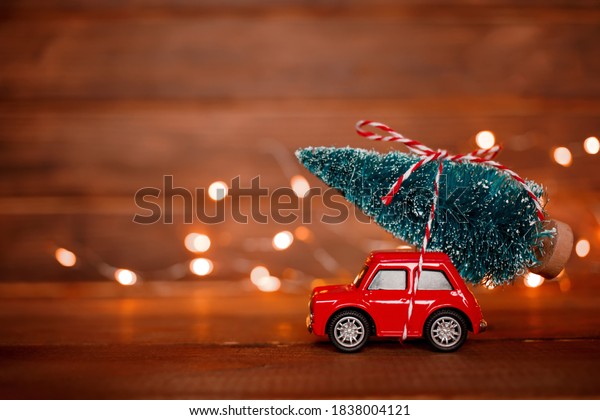 Miniature red car toy delivering christmas tree on\
wooden background with light. Christmas greeting card concept.\
Close up